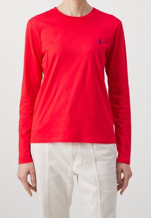 Personne portant une chemise rouge vif à manches longues avec un petit logo sombre sur la poitrine et un pantalon blanc sur un fond uni.