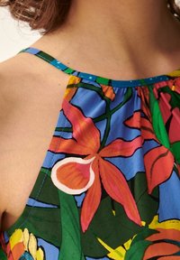 Top senza maniche a fantasia tropicale con fiori arancioni e rosa brillanti su uno sfondo blu, con scollatura arricciata e spalline verdi.