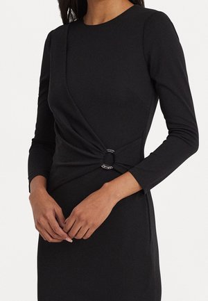 Femme portant une robe noire ajustée à manches longues avec un col rond et un détail torsadé maintenu par un anneau argenté à la taille.