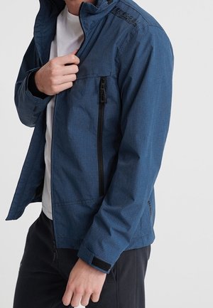 Mężczyzna w niebieskiej, teksturowanej kurtce z przednim zamkiem błyskawicznym i zamykana kieszenią na piersi, na białym T-shircie i ciemnych spodniach.