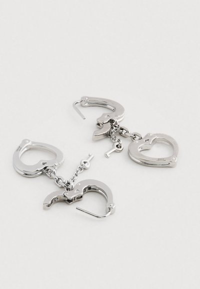 Strieborné náušnice v tvare putiek a sŕd, spojené reťazou, s malým detailom kľúča. Kovový a lesklý povrch.