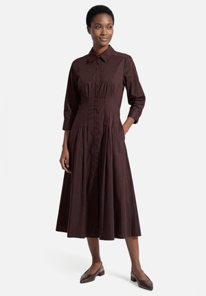 Frau steht, trägt ein dunkelbraunes, langärmeliges Midi-Kleid mit Kragen und Falten und passende niedrighackige spitze Schuhe.