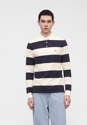 Jeune homme portant un polo à manches longues rayé horizontalement noir et blanc et un jean bleu clair, debout devant un fond uni.