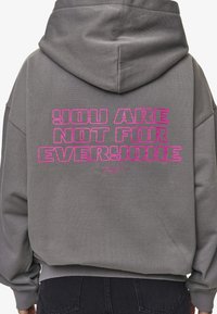 Hoodie grigio con testo rosa audace sul retro che recita "NON SEI PER TUTTI" con un messaggio più piccolo sotto. Tessuto morbido, vestibilità rilassata.