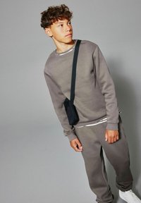 Adolescent avec des cheveux bouclés portant un sweat-shirt gris et un jogging, des baskets blanches et un sac bandoulière noir, regardant sur le côté devant un fond gris.