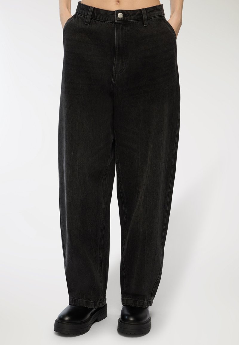 Persona che indossa jeans neri larghi con le mani nelle tasche e stivali chunky neri con plateau su uno sfondo neutro.