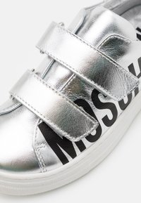 Sneaker sintetica argento con due cinturini in Velcro, con una stampa grafica nera sul lato e una suola in gomma bianca.