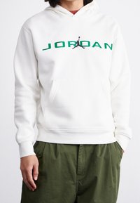 Felpa bianca con cappuccio caratterizzata da un logo "JORDAN" verde e un grafiche Jumpman nera, dotata di tasca frontale e polsini a costine. Abbinata a pantaloni verdi.
