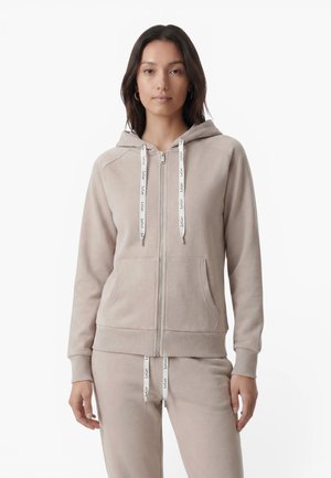Jeune femme portant un sweat à capuche beige zippé et un pantalon de survêtement assorti avec des cordons blancs, debout sur fond blanc.