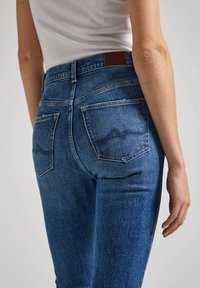 Tajta blå jeans med en slät denimtextur. Har två bakfickor med ett subtilt vågm�önster och en brun läderlapp på midjebanden.