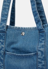 Blå denim totebag med to robuste håndtag, der har en frontlomme og et sølvmetalisk knapdetalje øverst.