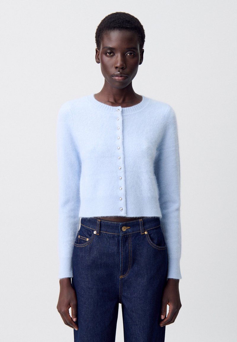Cardigan accorciato di un blu chiaro, realizzato in un materiale morbido e peloso, con scollatura rotonda e bottoni simili a perle lungo la parte anteriore, abbinato a jeans in denim scuri.