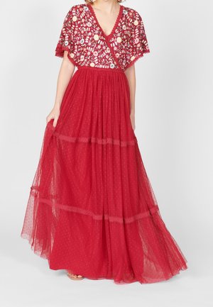 Kobieta ubrana w długą czerwoną suknię z górą z haftowanej koronki w kwiaty oraz przezroczystą spódnicę w kropki, lekko trzymająca jej brzeg.