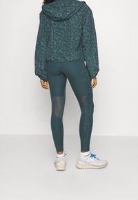Teal vzorovaná sportovní bunda s kapucí, spojená s odpovídajícími legínami s síťovými panely a hladkým povrchem, doplněná o světlé tenisky.