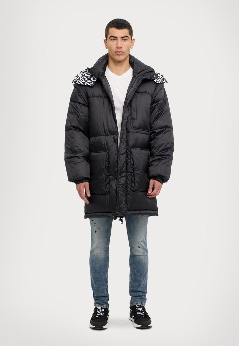 Chaqueta de plumas negra con cremallera en el frente, grandes bolsillos y una capucha con patrón. Combinada con unos vaqueros azul claros y zapatillas negras.