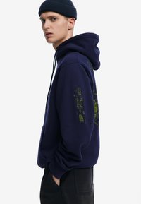 Marineblauwe hoodie met groene geborduurde ontwerpen op de mouwen en de voorkant. Bevat een kangoeroezak en een verstelbare trekkoordkap.