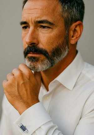 White dress shirt with a textured cuff. Silver cufflink with blue accents. Close-up of a man’s hand resting on his chin.
