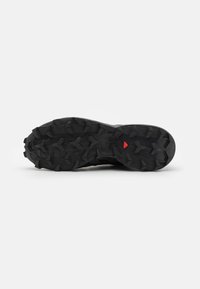 Semelle de chaussure noire robuste avec des crampons profonds multidirectionnels et un petit détail triangulaire rouge près du centre.