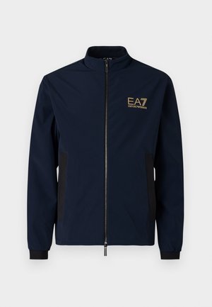 Marineblå vindjakke med høj krave, lynlås-lukning, sorte sidepaneler og et guld EA7-logo på venstre bryst. Glat, letvægtsmateriale.