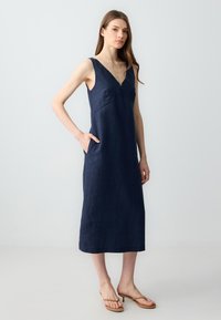 Marineblaues Leinenkleid mit tiefem V-Ausschnitt, Schulterträgern und seitlichen Taschen; fällt unter das Knie mit einer geraden Silhouette.