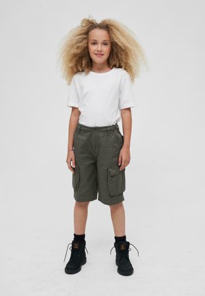 Witte shirt met korte mouwen, groene cargoshorts met meerdere zakken, zwarte enkellaarzen en donkere sokken. Effen achtergrond.