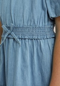 Osvetljeno modrije denim obleka ima elastičen pas s pritrjenim lokom in nabranim materialom, kar poudarja ohlapen, tekoč kroj.