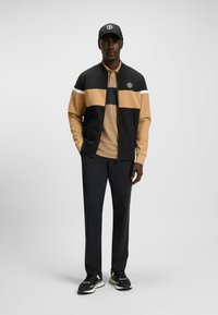 Sport jacket in black and beige with a zip front, paired with a tan polo shirt and black trousers. Features logo on the cap.