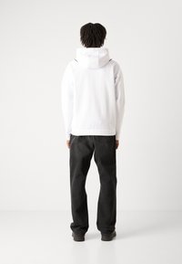 Sweatshirt à capuche blanche avec une texture lisse, agrémenté d'accents noirs, associé à un jean gris foncé. La coupe est ample et décontractée.
