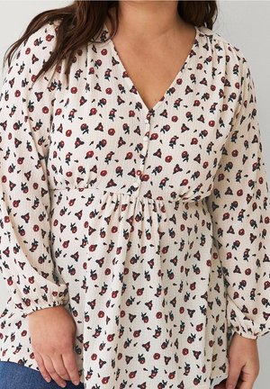 Vrouw draagt een crèmekleurige blouse met lange mouwen en een rood-zwarte bloemenprint, met knopen aan de voorkant, gecombineerd met een blauwe spijkerbroek.