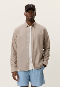 Ανοιχτό μπεζ πουκάμισο με κουμπιά, υφαντή υφή, λευκό μπλουζάκι από μέσα, σε συνδυασμό με ανοιχτό μπλε σορτς με σχέδια και σκουρόχρωμο καπέλο με λογότυπο.