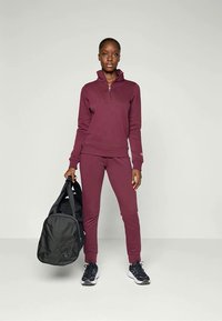 L'outfit sportivo color bordeaux include una felpa con zip e pantaloni aderenti. Modella che tiene una borsa da palestra nera. Sneakers nere completano l'outfit.