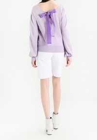 Levandulový pletený svetr s výstřihem do V na zádech a fialovou mašlí, v kombinaci s bílými šortkami a kotníkovými botami. Hladká textura, volné rukávy.