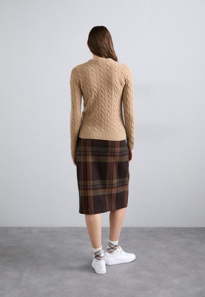 Beige cable-knit sweater with a fitted design, paired with a knee-length plaid skirt featuring dark brown, green, and red stripes. White sneakers.