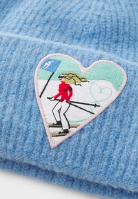 Light blue knitted hat featuring a heart-shaped patch with an embroidered skier in red, against a green background with a blue flag.