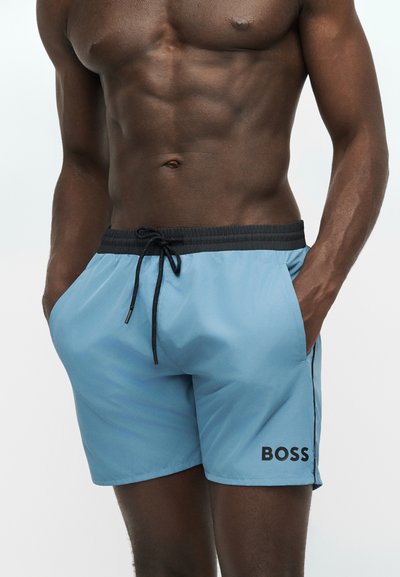 Homme musclé portant un short de bain bleu clair avec une ceinture noire et le logo "BOSS", debout les mains dans les poches sur un fond uni.