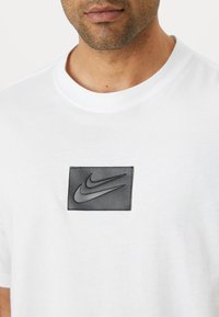 Witte katoenen t-shirt met een zwart leren patch met een reliëf Nike-logo. Eenvoudig crewneck ontwerp met korte mouwen.