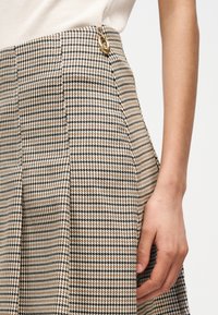 Χέρι γυναίκας ακουμπισμένο στο ισχίο της, φοράει φούστα με σχέδιο κυνόδοντα σε μπεζ και μαύρο, με χρυσό κυκλικό κούμπωμα στη μέση.