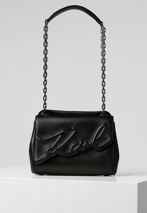 Bolso de mano de cuero negro con correa de cadena acolchada, cierre de solapa y detalle de escritura en relieve en la parte delantera que dice "Karl."