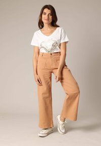 Witte V-hals t-shirt met een goudkleurige zebra hartafbeelding, gecombineerd met hoge taille, wijde lichte tan broek en witte sneakers.