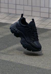 Schwarze Leder-Sneaker mit dicker Gummisohle, Schnürdesign, abgerundeten Kanten und strukturierten Paneelen. Über einer strukturierten Oberfläche schwebend.
