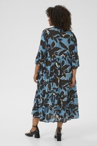 Kobieta z kręconymi włosami, ubrana w niebieską i czarną sukienkę midi w kwiaty oraz czarne buty na słupku, stojąca tyłem na jednolitym tle.