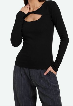 Femme portant un haut noir ajusté à manches longues avec une découpe près de l'encolure et un pantalon gris rayé ample, main dans la poche.