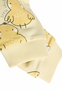 Dzeltena auduma, uz kura attēloti zīmēti cāļi, ar ribinātiem piedurknēm. Mīksta tekstūra ar vieglu, pasteļtoņu krāsu.