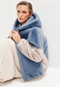 Cappotto con cappuccio in pelliccia sintetica, con corpo blu e maniche beige, caratterizzato da una texture morbida e una zip argento. Il modello posa seduto.