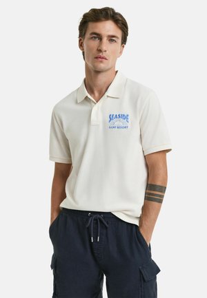 Kerok polo shirt dengan grafik biru "Seaside", lengan pendek, placket dua tombol, dipadukan dengan celana pendek navy. Tekstur lembut, desain kasual.