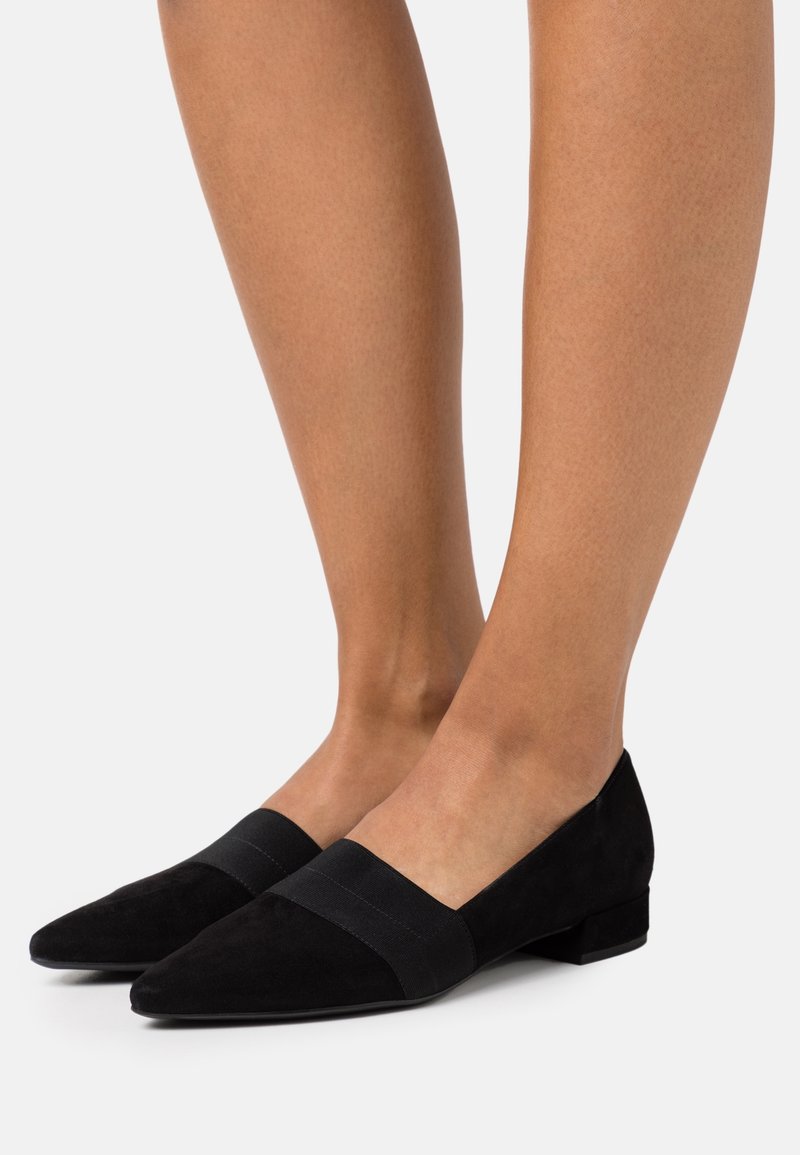 Jambes portant des chaussures plates en daim noir à bout pointu avec un détail en bande élastique sur un fond blanc.