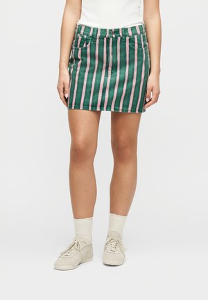 Γυναίκα που φοράει πράσινη και ροζ μίνι φούστα με κάθετες ρίγες, λευκές κάλτσες, λευκά αθλητικά παπούτσια και άσπρο τοπ, μπροστά σε απλό φόντο.