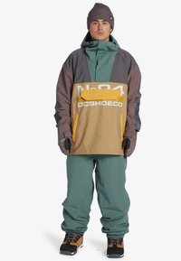 Bunte Snowboardjacke in Teal, Braun und Gelb, mit großer Fronttasche und Logo; kombiniert mit passenden Hosen und Handschuhen.