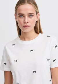 White cotton t-shirt with black bow embroidery scattered throughout. Features a round neckline and short sleeves, with a smooth texture.
