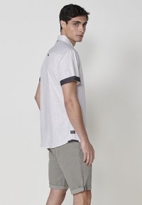 Camisa de botones de manga corta en tela texturizada gris claro con acentos en puños azul oscuro. Combina con pantalones cortos grises remangados.